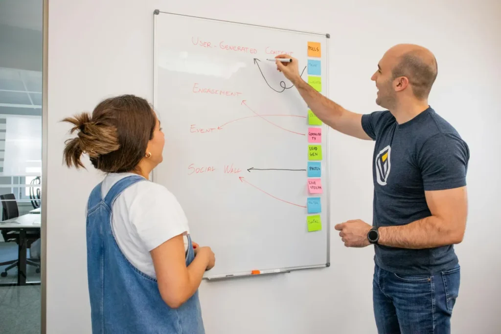 Dua rekan kerja berdiskusi strategi marketing di depan papan tulis dalam suasana kantor modern